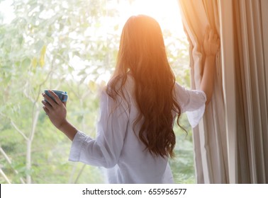 Wake Up :Woman In The Morning Holding A Cup Of Tea Or Coffee And Looking At The Sunrise Standing Near The Window In Her Home