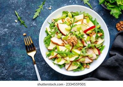 Waldorf salad with fresh apple, celery, lettuce, grilled chicken fillet, arugula and walnuts on plate, blue table background, top view