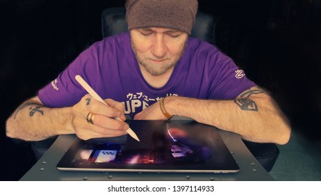 Wales, UK / May 14th 2019 : bearded man with tattoos editing photos using Affinity Photo on an iPad Pro with an Apple Pencil.