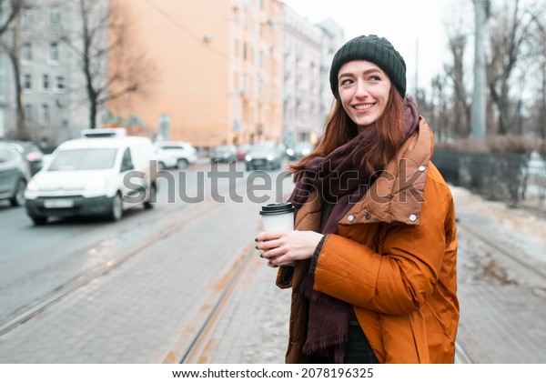 Walk around the city in the cold season\
with a cup of coffee. Orange long jacket, scarf and warm hat. Looks\
away. Blurry image of the city and\
cars.