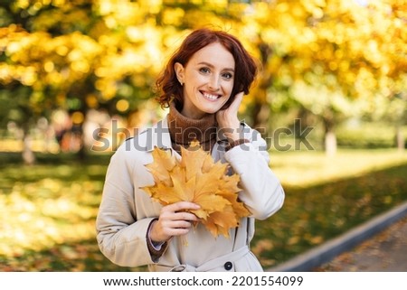 Walk in city. Cheerful caucasian young redhead female in raincoat hold yellow leaf, enjoy freedom and warm weather in park in autumn. Ad and offer, active lifestyle and health care during covid-19