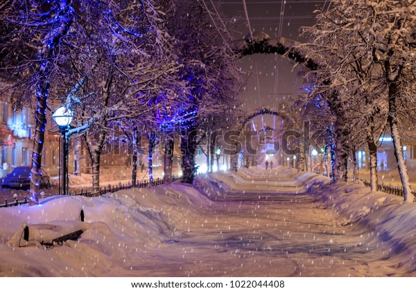 Walk of the city\
at night in winter. Alley with lights, trees, garlands covered with\
snow. Cityscape. Snowfall.