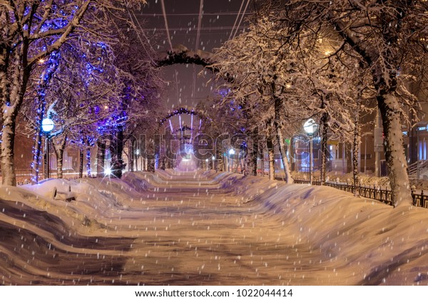 Walk of the city\
at night in winter. Alley with lights, trees, garlands covered with\
snow. Cityscape. Snowfall.