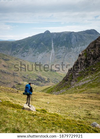A walker wearing a backpack looks towards the Langdale valley and Pike of Stickle beyond Black Wars, cliffs below Pike o' Blisco, a fell (or mountain) in the Lake District, Cumbria, England.