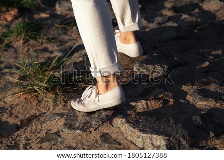 Walking along the beach. Female legs in white trousers and shoes go over stones on a sandy beach close up
