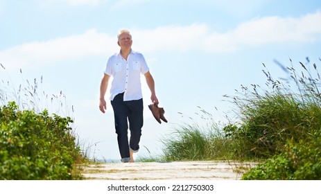 Walking Barefoot Through The Park. A Senior Man Walking Through A Park With His Shoes In Hand.