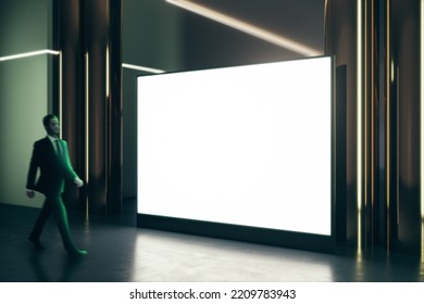 Walking Businessman By Blank White Illuminated Big Screen With Space For Your Logo Or Text In Dark Empty Room With Glossy Floor, Green Wall And Golden Pillars, Mock Up