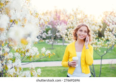 Walking By Spring Park. Attractive Young Woman Talking By Phone And Holding Cup Of Coffee Outdoors.