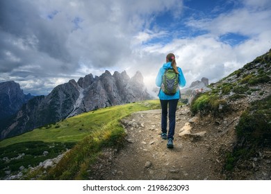 Walking girl with backpack on the trail in mountains at sunset in summer. Beautiful landscape with young woman, high rocks, path, green grass, cloudy sky in Dolomites, Italy. Adventure and hiking