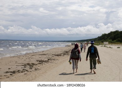 Walking With Social Distancing. Couple Walks Apart From Other People By Sea Coast Or Ocean Edge Along The Sandy Beach. Background Landscape Of Travel, Tourism, Leisure, Family And Active Lifestyle.