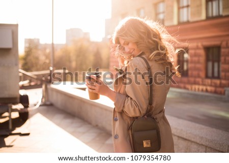 Walking is a spiritual journey and a reflection of living! Warm and sunny professional photo of the young smart model smiling and drinking coffee on the street, while smoothing her wavy hair.
