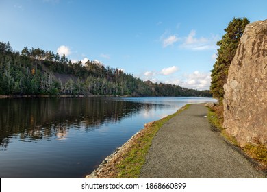 A walking trail of crushed stone and gravel along a wide river. The trail has water on one side and a rock face cliff on the other side. The stream is calm and reflects the mountain covered in trees