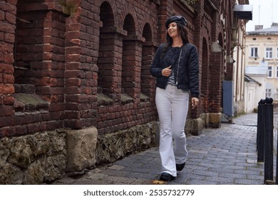 Walking woman, rain photography, fashionable outfit. Dressed in black jacket and white trousers, fashionable woman casually smiles as she walks by brick wall on rainy day. - Powered by Shutterstock