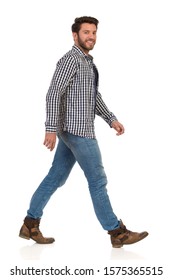 Walking young man in boots, jeans and lumberjack shirt, smiling and looking at camera. Side view. Full length studio shot isolated on white.