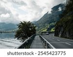 Walkway along the northern bank of Alpnachersee mountain lake. POV: walking on narrow boardwalk aside paved road by the edge of the water. Swiss Prealps covered in clouds in the background.