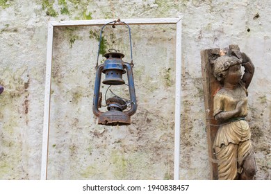 The Wall Of The Abandoned Garden. Kerosene Lamp And Wooden Child Statue Hanging On It.