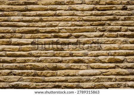 A wall is built using yellow brown stones stacked in horizontal lines. Sunlight highlights the texture and pattern of the stones