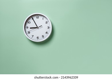 Wall clock hanging on the pale green wall with copy space. Round white clock with black hands. Five minutes to nine. Time measuring, hour and minutes concepts. Time control. Working hours. Front view.
