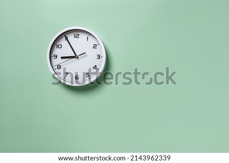 Wall clock hanging on the pale green wall with copy space. Round white clock with black hands. Five minutes to nine. Time measuring, hour and minutes concepts. Time control. Working hours. Front view.