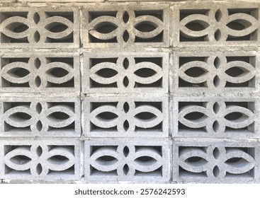 A wall composed of light gray concrete blocks. The blocks are arranged in a regular grid pattern.  Each block features a repeating, symmetrical design of curved, interlocking shapes creating a floral.