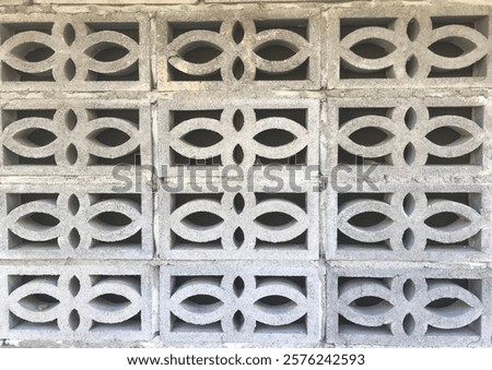 A wall composed of light gray concrete blocks. The blocks are arranged in a regular grid pattern.  Each block features a repeating, symmetrical design of curved, interlocking shapes creating a floral.