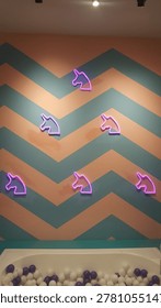 A wall decorated with a bold chevron pattern in pastel peach and teal features multiple glowing neon unicorn silhouettes emitting a soft magical aura, creating a whimsical and enchanting. 