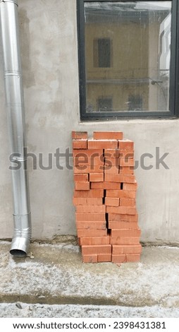 the wall of the house has a window and a drainpipe, bricks are stacked next to it. The window reflects the neighboring building