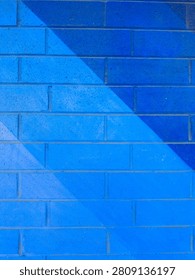 A wall made of bricks is painted in several shades of blue. Light hits the surface, creating different tones. This scene is set in a city during midday.
