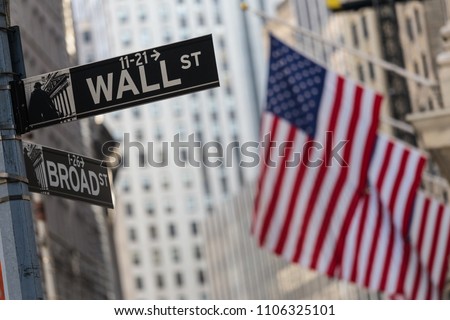 Wall street sign in New York City with American flags and New York Stock Exchange in background.