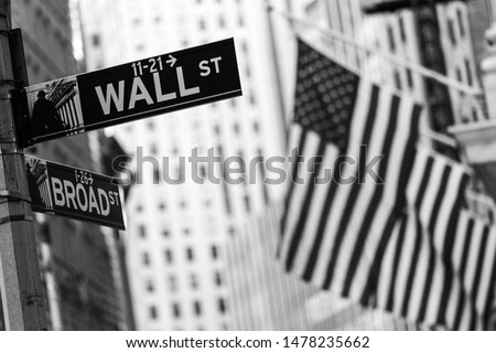 Wall street sign in New York City with American flags and New York Stock Exchange in background. Black and white image.