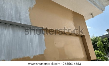 A wall surface is shown during a painting or renovation process. Sections are covered in a light gray primer or undercoat, while the majority of the wall features a warm, light brown topcoat, indicati