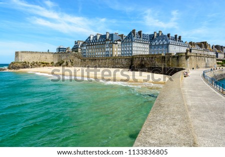 The walled city of Saint Malo in Brittany, France, with granite residential buildings sticking out above the rampart and people sunbathing on the Mole beach at the foot of the high wall.