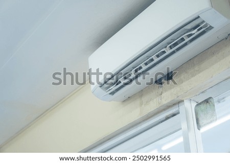 A wall-mounted air conditioning unit installed in a room. The unit is white and features vents for airflow. The wall shows signs of wear, with some discoloration and peeling paint.