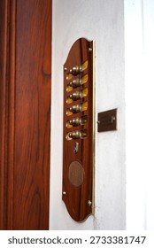 Wall-mounted interior bell panel made of polished wood with a vertical arrangement of small brass bells and mechanical components. The handcrafted appearance and warm materials suggest a traditional signaling or call system used in historic buildings, residences, or institutional interiors. Each bell is individually mounted, indicating separate lines, rooms, or functions once connected to a mechanical or wired alert mechanism. The smooth wooden backing contrasts with the metallic shine of the bells, highlighting craftsmanship, durability, and decorative function combined with practical use. Installed beside a doorway, the panel reflects an earlier era of communication technology, where sound-based signals replaced electronic indicators. This image conveys themes of heritage engineering, vintage technology, craftsmanship, and historical infrastructure. It is well suited for concepts related to architectural details, antique interiors, traditional signaling systems, and the evolution of communication tools. The documentary composition and material clarity make it ideal for editorial storytelling, historical references, interior design features, and visual narratives about functional objects from the pre-digital age.