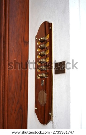 Wall-mounted interior bell panel made of polished wood with a vertical arrangement of small brass bells and mechanical components. The handcrafted appearance and warm materials suggest a traditional signaling or call system used in historic buildings, residences, or institutional interiors. Each bell is individually mounted, indicating separate lines, rooms, or functions once connected to a mechanical or wired alert mechanism. The smooth wooden backing contrasts with the metallic shine of the bells, highlighting craftsmanship, durability, and decorative function combined with practical use. Installed beside a doorway, the panel reflects an earlier era of communication technology, where sound-based signals replaced electronic indicators. This image conveys themes of heritage engineering, vintage technology, craftsmanship, and historical infrastructure. It is well suited for concepts related to architectural details, antique interiors, traditional signaling systems, and the evolution of communication tools. The documentary composition and material clarity make it ideal for editorial storytelling, historical references, interior design features, and visual narratives about functional objects from the pre-digital age.