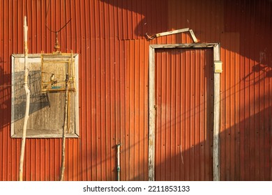 The Walls Of The House Are Made Of Galvanized Sheets. Brown And Orange It Is Plastered With Sunlight And Has White Doors, Windows And A Wooden Birdcage. There Is A Bird In The Cage