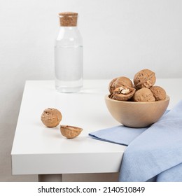 Walnuts lie in a wooden bowl standing on a white table. There is a carafe of water in the background, and a blue towel is lying next to the nuts.