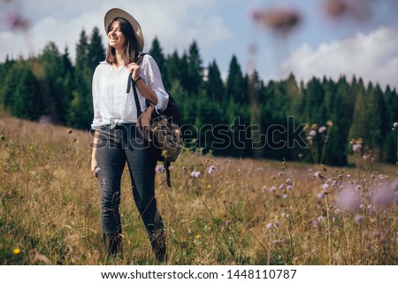 Wanderlust and travel concept. Hipster girl with backpack traveling in sunny mountains, walking in  wildflower meadow. Stylish happy woman in hat exploring and hiking on top of mountain.