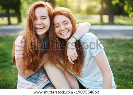 Wanna hug her forever. Portrait of happy and carefree two twin sisters with natural red hair and freckles, laughing out loud and cuddling, fooling around while resting in park on fresh green grass.