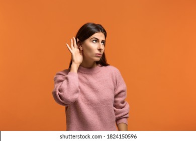 I want to hear everything. I want to know. Curious woman in pink sweater holds and near ear and to overhear interesting information against orange background indoor