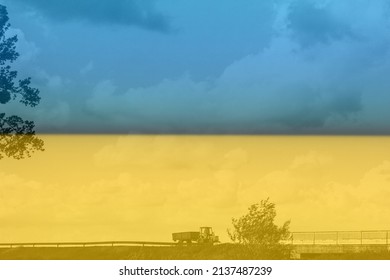 War Ukraine. Ukrainian Flag. A Red Tractor With A Green Trailer Drives Onto A Bridge In The Countryside In Windy Weather. You Can See The Grass, Trees, Clouds.