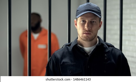 Warden Looking At Camera In Prison, African American Criminal On Background