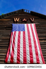 Wardensville, West Virginia/USA- May 25, 2018: A Vertical Image Of An Over-sized American Flag Hanging Down The Front Of A Vintage And Weathered Brown Barn.                        