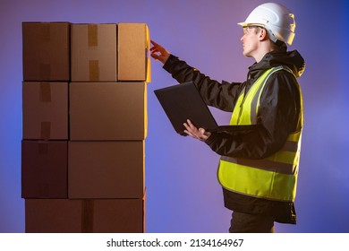 Warehouse Accounting. Man Counts Number Of Boxes. Man Dressed As Warehouse Worker. Guy Is Engaged In Warehouse Accounting. Man In Work Uniform Holds Laptop. Fulfillment Company Employee On Dark.