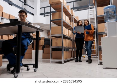 Warehouse delivery managers using order processing system on laptop and checking data on clipboard. Post office storehouse asian women employees managing inventory control