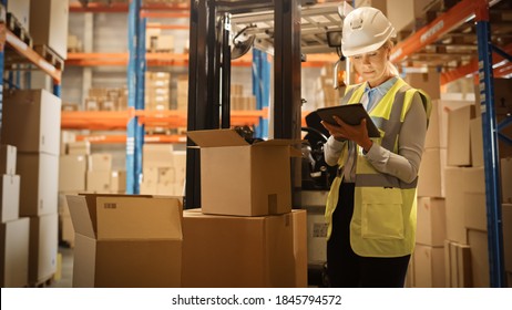 In Warehouse Female Manager Wearing Hardhat Standing Beside Forklift Uses Digital Tablet For Inventory, Checking Inside Of Cardboard Box Package. Distribution, Delivery Center, Shelves With Packages