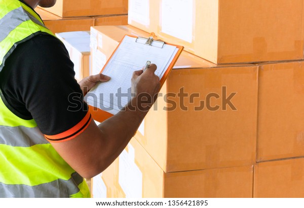 Warehouse inventory\
and management, Young warehouse worker is inspecting the products,\
stack of cardboard\
boxes.