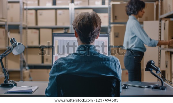 Warehouse Inventory Manager Talks to a Clerk\
Working with a Spreadsheet on a Personal Computer while Sitting at\
His Desk. In the Background Shelves Full of Cardboard Box Packages\
Ready For Shipping.