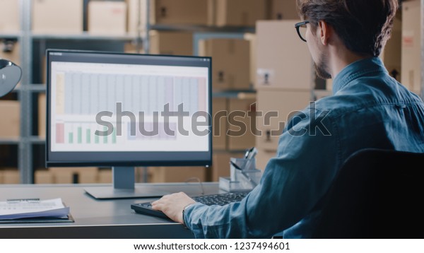 Warehouse\
Inventory Manager Works with a Spreadsheet on a Personal Computer\
while Sitting at His Desk. In the Background Shelves Full of\
Cardboard Box Packages Ready For\
Shipping.