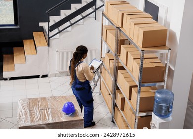 Warehouse manager checking documentation on clipboard and inspecting parcels. African american woman storehouse supervisor overseeing packages dispatching operations top view
